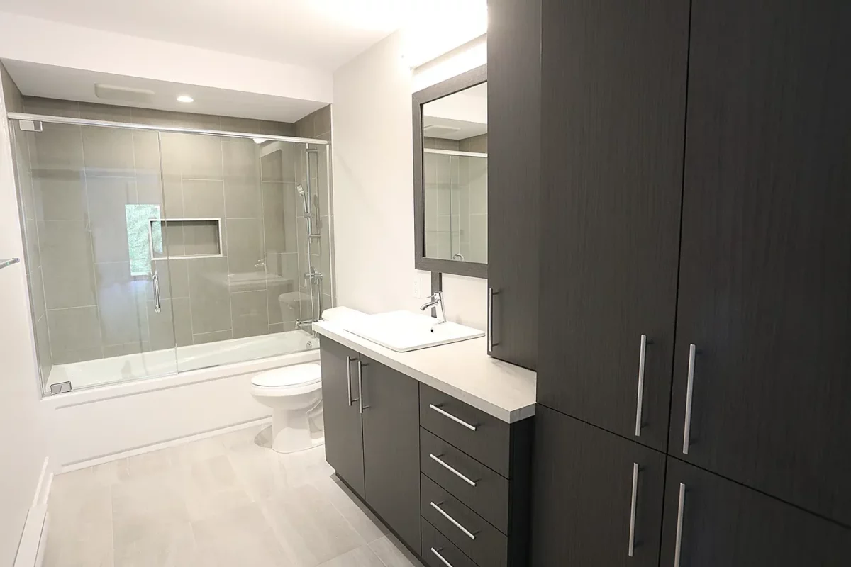 Salle de bain moderne avec douche vitrée, armoires brunes et vanité blanche – projet multi-résidentiel Joliette par BGC