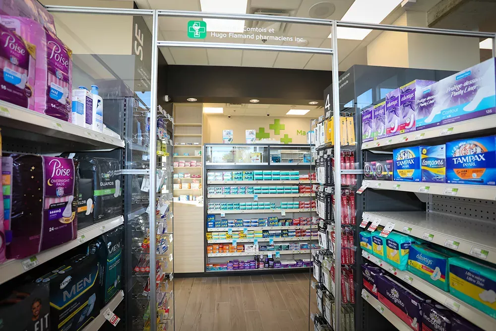 Allée de pharmacie avec étalages de produits et croix verte en arrière-plan – Familiprix Joliette par Gestion BGC