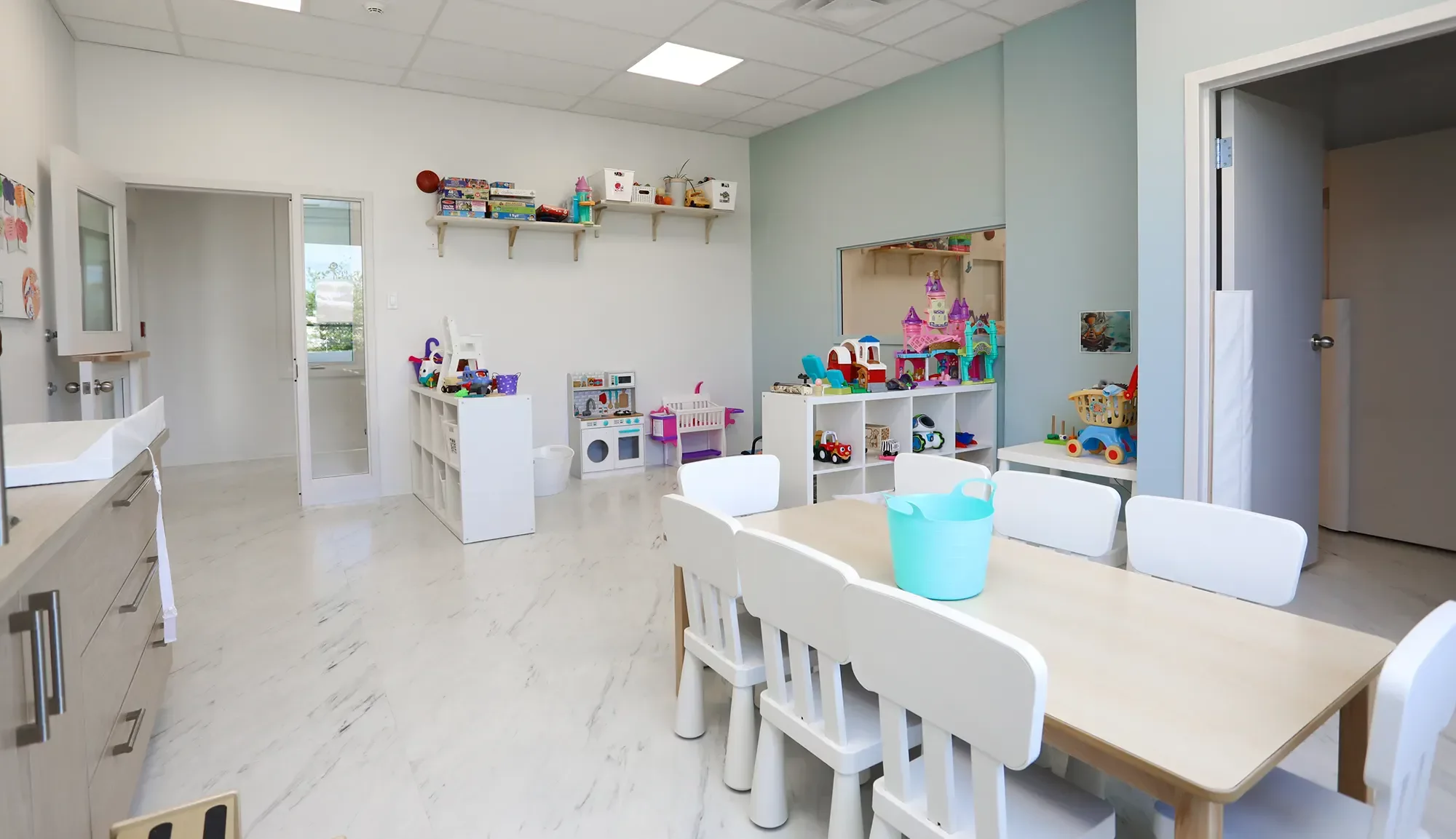 Salle de jeux avec fenêtres d'observation entre les locaux, dessins d'enfants et étagères – Garderie Maxi-Mousse